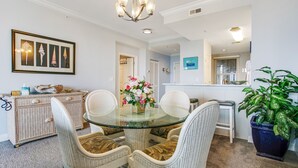 A dining area with a glass-top table, wicker chairs, a floral centerpiece, a white console table with decorative items, and a potted plant. - Luxe, Updated, Oceanfront Condo with Ground-Floor Patio, WiFi & Shared Pool (Fernandina Beach)