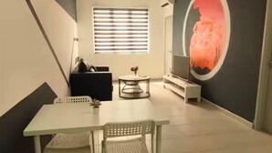 A dining area with a white table and chairs, a black sofa, a small round table with a vase, and a large TV on a stand. - JetPad Suites - Near KLIA (Dengkil)