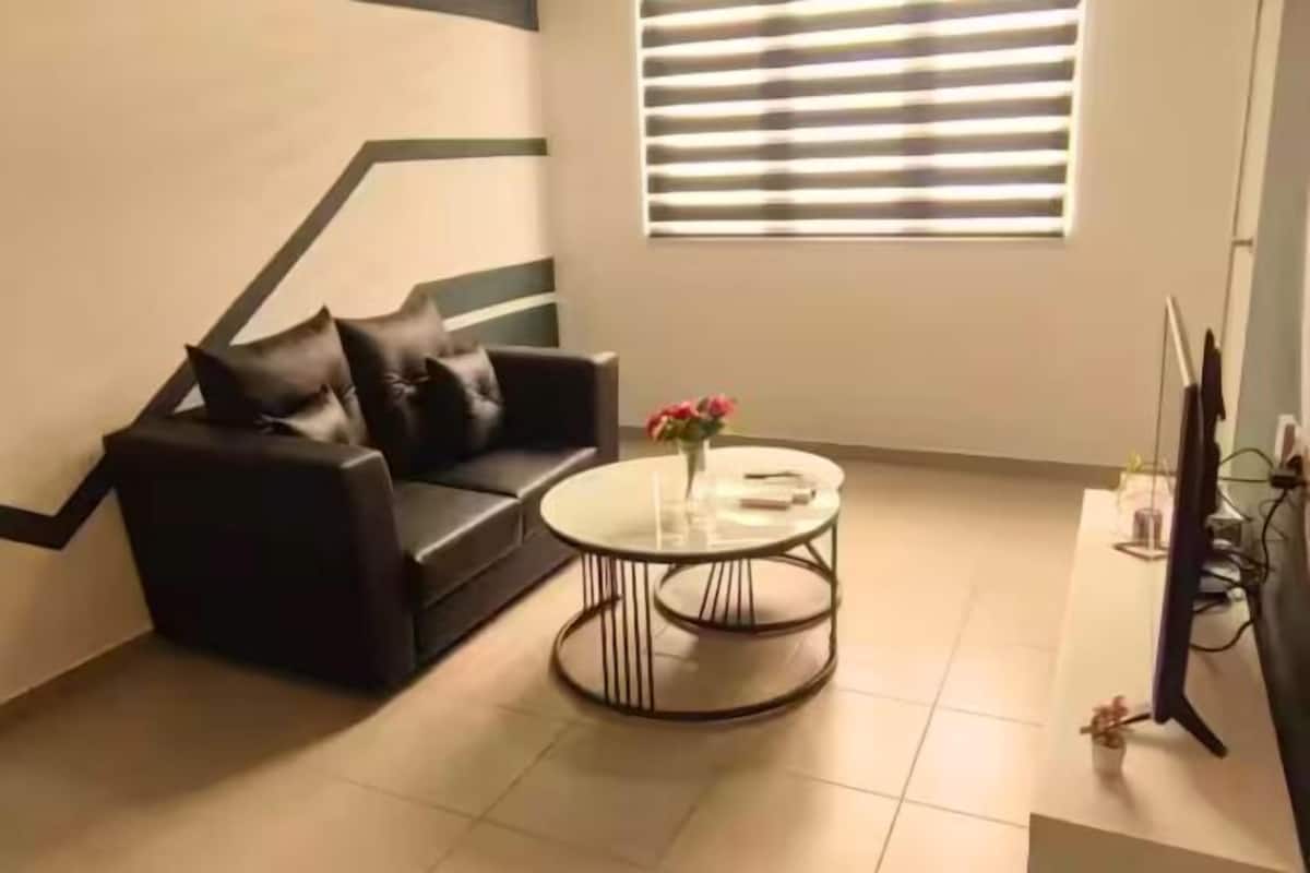 Un salon avec un canapé en cuir noir, une table basse en verre et une fenêtre avec des stores.