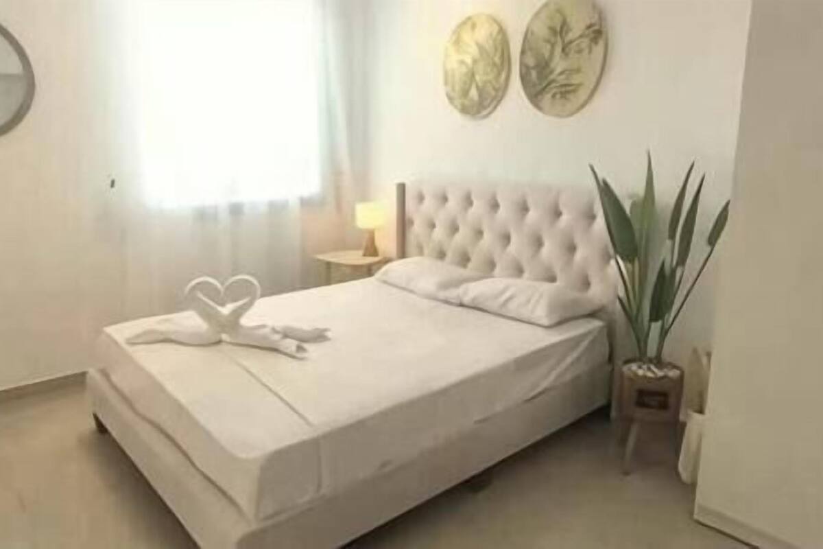 A neatly made bed with white linens and heart-shaped towels, a round mirror on the wall, and a potted plant beside the bed.