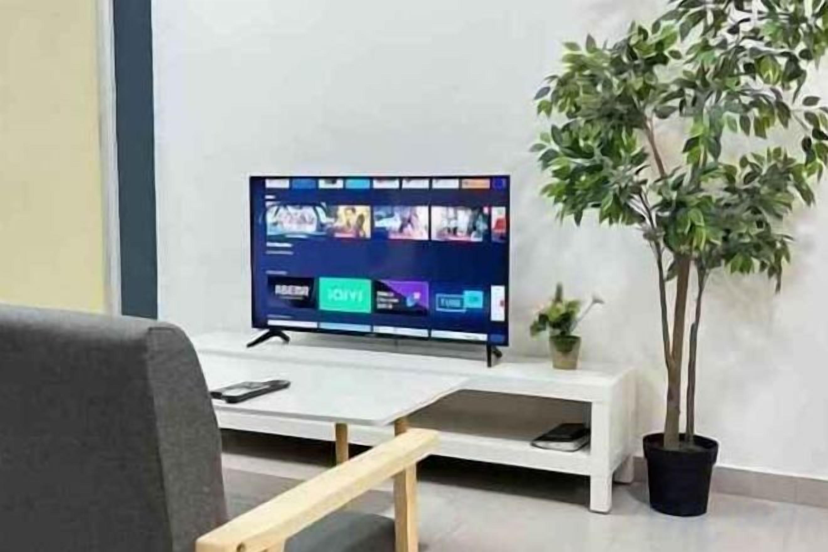 A flat-screen TV on a stand, a potted plant, and a white bench in a room with a plain wall.