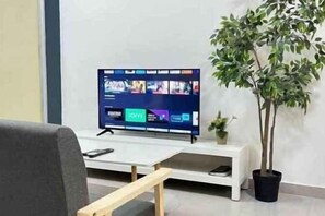 A flat-screen TV on a stand, a potted plant, and a white bench in a room with a plain wall. - JetPad Suites - Near KLIA (Dengkil)