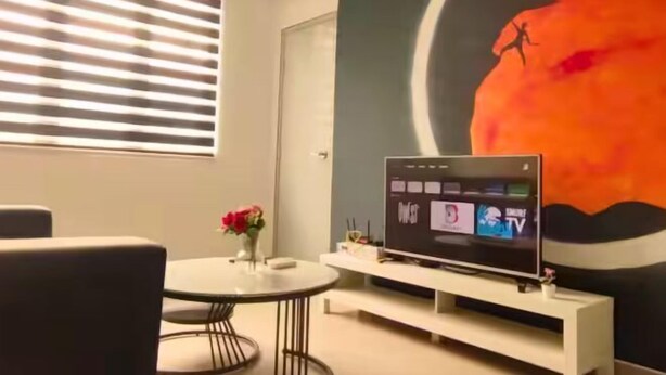 A room with a round table, a flat-screen TV on a white stand, a vase with flowers, and a large wall mural. - JetPad Suites - Near KLIA (Dengkil)