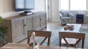 A living room with a TV on a wooden cabinet, a patterned armchair, and a coffee table. - Beautiful Condo with Gorgeous Ocean Views from Balcony + Shared Pool & Free WiFi (Fernandina Beach)