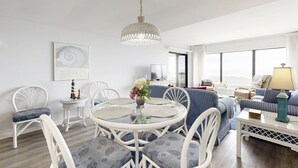 Un salon moderne avec une table ronde en verre, des chaises blanches, un canapé à rayures bleues et une grande fenêtre donnant sur un paysage agréable.
