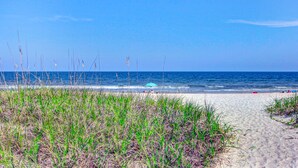 Une plage avec un sentier de sable menant à l’eau, de l’herbe au premier plan et un ciel bleu dégagé.