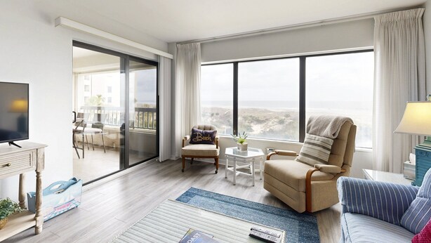 Une chambre d’hôtel moderne dotée d’une grande porte-fenêtre coulissante donnant sur un balcon, d’un coin salon confortable avec un canapé et des fauteuils, d’un téléviseur à écran plat et d’une petite table agrémentée d’un vase.
