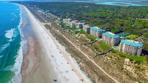 Vue aérienne d’une zone côtière avec une plage, un sentier pédestre et plusieurs immeubles à étages dotés de toits bleus.