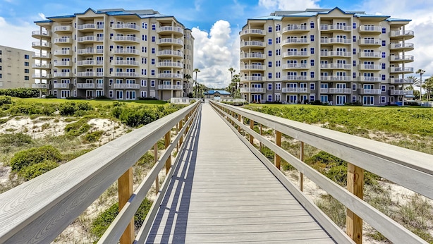 Une promenade en bois mène à un immeuble d’appartements situé en bord de mer, doté de nombreux balcons et offrant une vue sur un ciel dégagé.