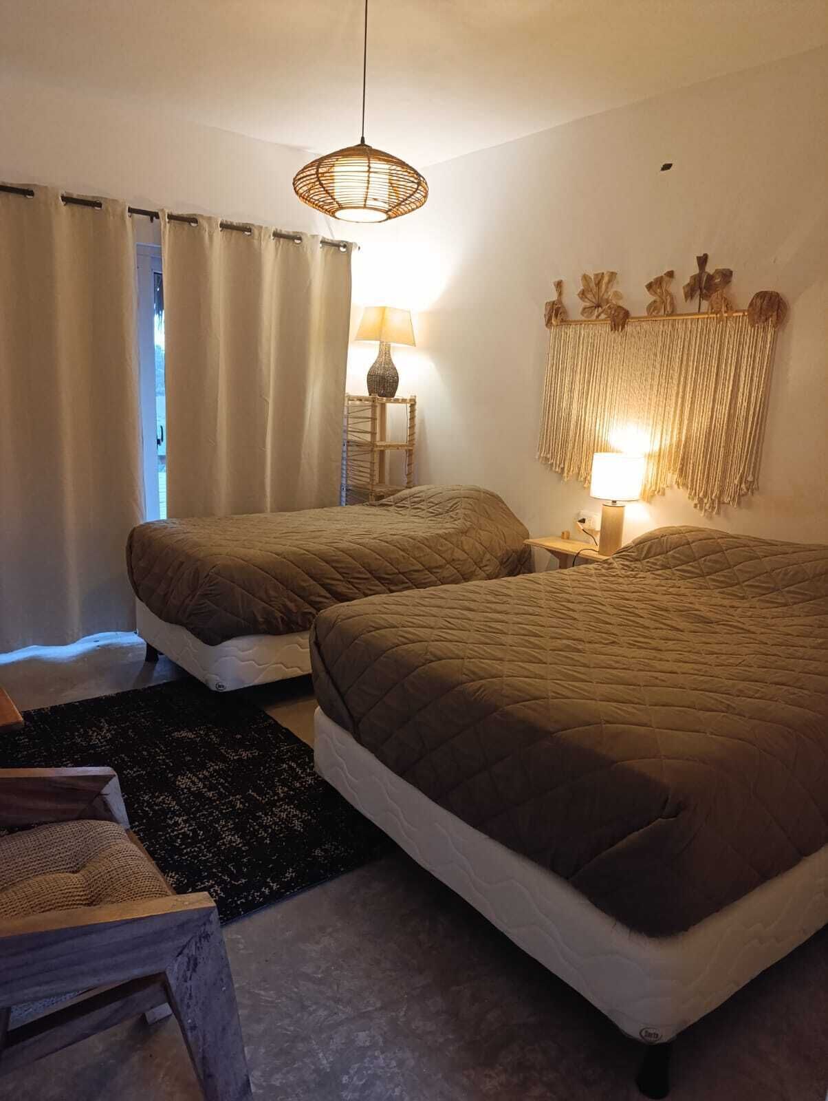 A bedroom with two beds, a bedside table with a lamp, a hanging light fixture, and a wall-mounted macrame decoration.