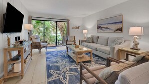 A living room with a TV, two sofas, a coffee table, and a large window with a view of greenery. - Dog-friendly Beachwood home in Omni Plantation with pool & beach access (Fernandina Beach)