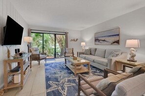 A living room with a TV, two sofas, a coffee table, and a large window with a view of greenery.