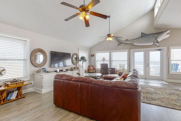 A spacious living room with a brown leather sofa, a ceiling fan, a mounted shark decoration, a flat-screen TV, and a large window providing a view of the outdoors. - Spacious Beachfront Home w/Deck, Balcony, Central A/C, Washer/Dryer, and WiFi (Fernandina Beach)