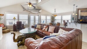 A spacious living room with a large brown leather sofa, a glass coffee table, and a ceiling fan. There are multiple windows with blinds, a kitchen area in the background, and decorative items like a shark and elephant sculpture. - Spacious Beachfront Home w/Deck, Balcony, Central A/C, Washer/Dryer, and WiFi (Fernandina Beach)