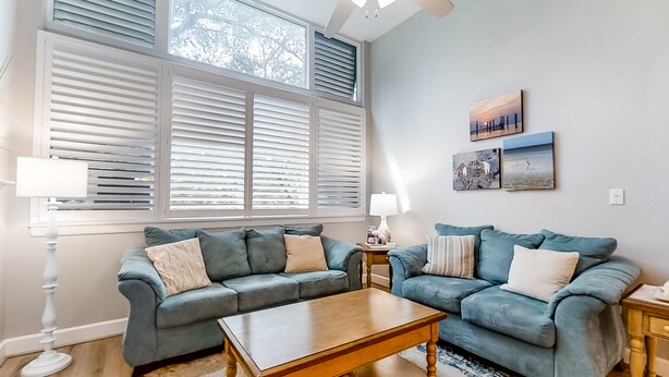 A living room with two blue sofas, a wooden coffee table, and a large window with blinds. - Renovated, dog-friendly condo near beach with balcony, pool & tennis (Fernandina Beach)