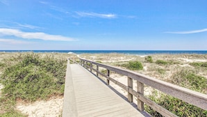 Une promenade en bois qui mène à une plage bordée de dunes de sable et d’une végétation clairsemée.