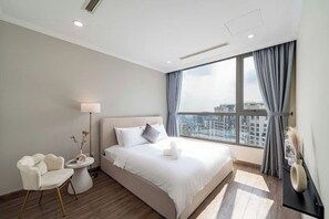 Habitación de hotel moderna con una cama grande, una mesita de noche, una silla, una lámpara de pie y vistas a la ciudad a través de un ventanal amplio.