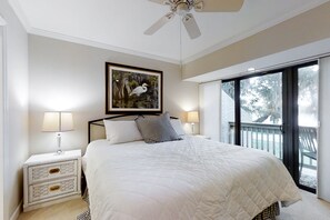 A bedroom with a large bed, two bedside lamps, a ceiling fan, a framed picture of a bird, and a view through a sliding glass door. - Updated & lovely villa with AC - steps from pool & beach (Fernandina Beach)