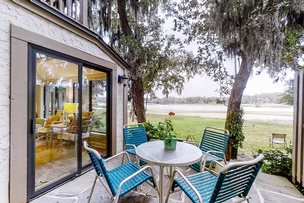 A patio with a round table and blue chairs, overlooking a grassy area with trees and a distant view. - Updated & lovely villa with AC - steps from pool & beach (Fernandina Beach)