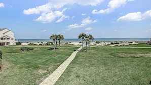 Une pelouse bien entretenue, avec un chemin pavé menant à une plage, des palmiers et une maison en arrière-plan.