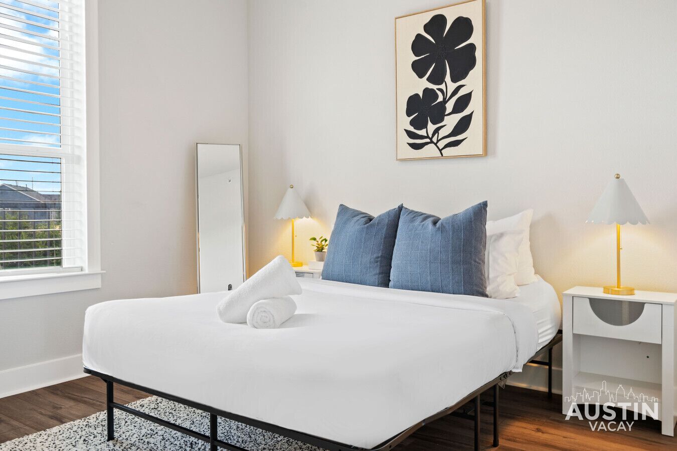 Ein modernes Hotelzimmer mit einem großen Bett, zwei Nachttischen mit Lampen, einem großen Spiegel und einem an der Wand befestigten Kunstwerk.