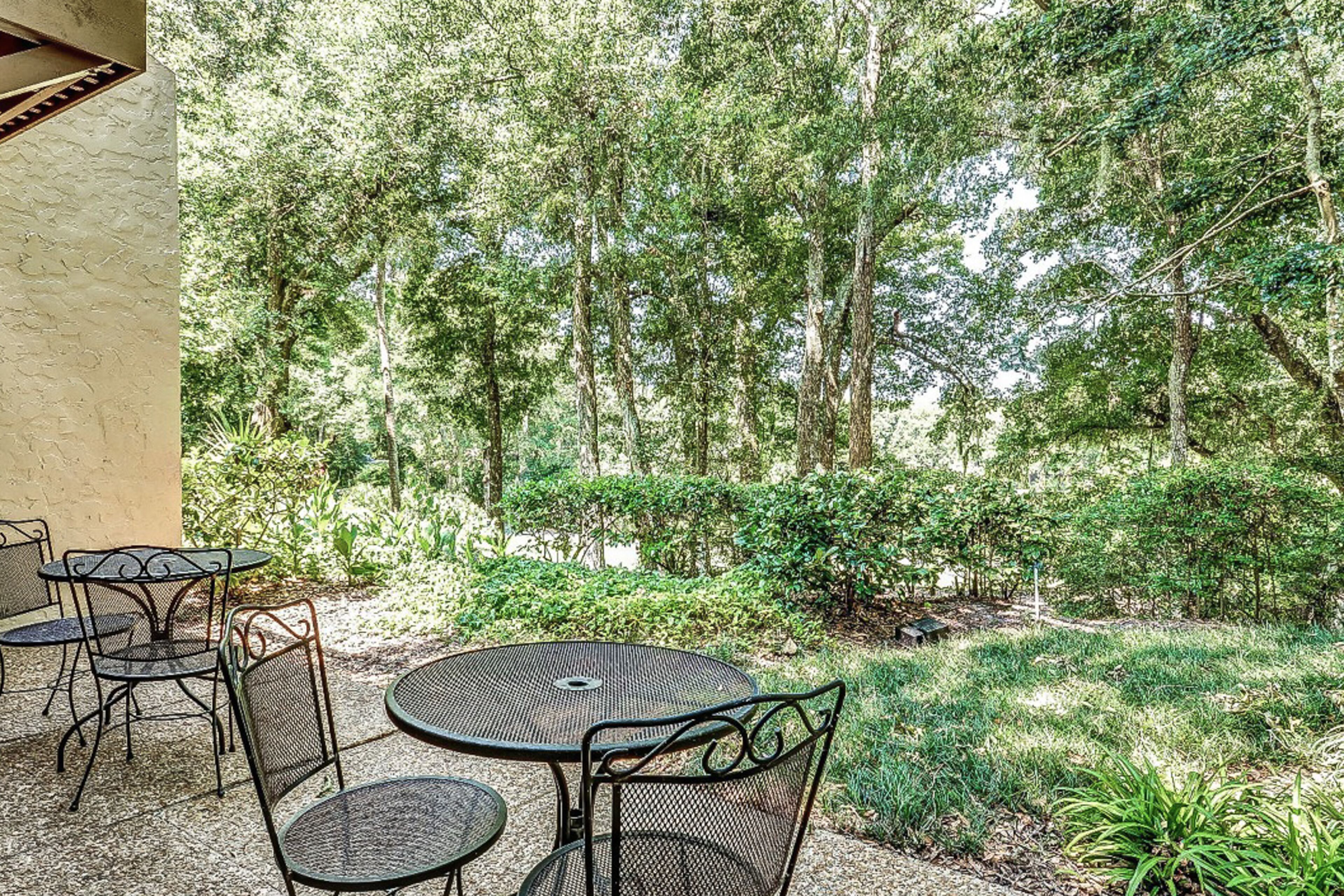 An outdoor patio with round tables and chairs, surrounded by dense greenery and trees.