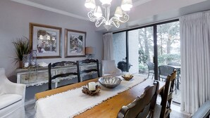 Uma área de refeições com mesa de madeira, cadeiras brancas, um candelabro, quadros enquadrados e vista para o exterior através de grandes janelas.