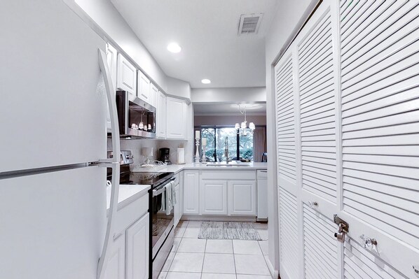 A narrow kitchen with white cabinetry, a built-in oven, and a window with a view of trees.