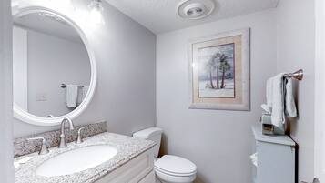 Een badkamer met een witte wastafel, toilet en een spiegel met verlichting. Er hangt een lijst met een foto aan de muur.