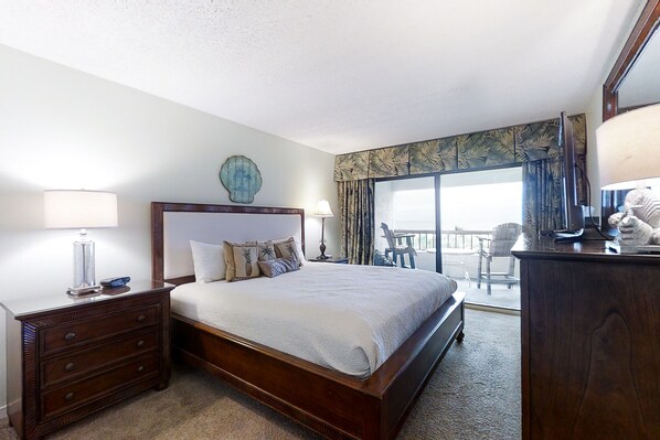 A hotel room with a large bed, a wooden nightstand with a lamp, a dresser, and a balcony with a view.
