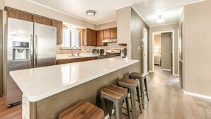 A modern kitchen with a central island, wooden bar stools, stainless steel appliances, and a view into the adjacent living area. - Juniper Retreat (Ruidoso)
