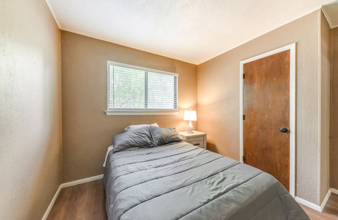 A bedroom with a bed, a nightstand, a lamp, a window with blinds, and a door.