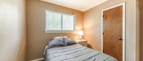 A bedroom with a bed, a nightstand, a lamp, a window with blinds, and a door.