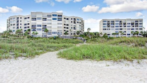 A beachfront hotel with multiple floors, a sandy beach in the foreground, and palm trees.