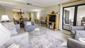 A spacious living room with a sofa, armchairs, a coffee table, and a television. There is a dining area in the background with a table and chairs.