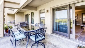 Une terrasse extérieure avec une table et des chaises à plateau en verre, une plante en pot et une vue sur l’intérieur à travers une porte-fenêtre coulissante.