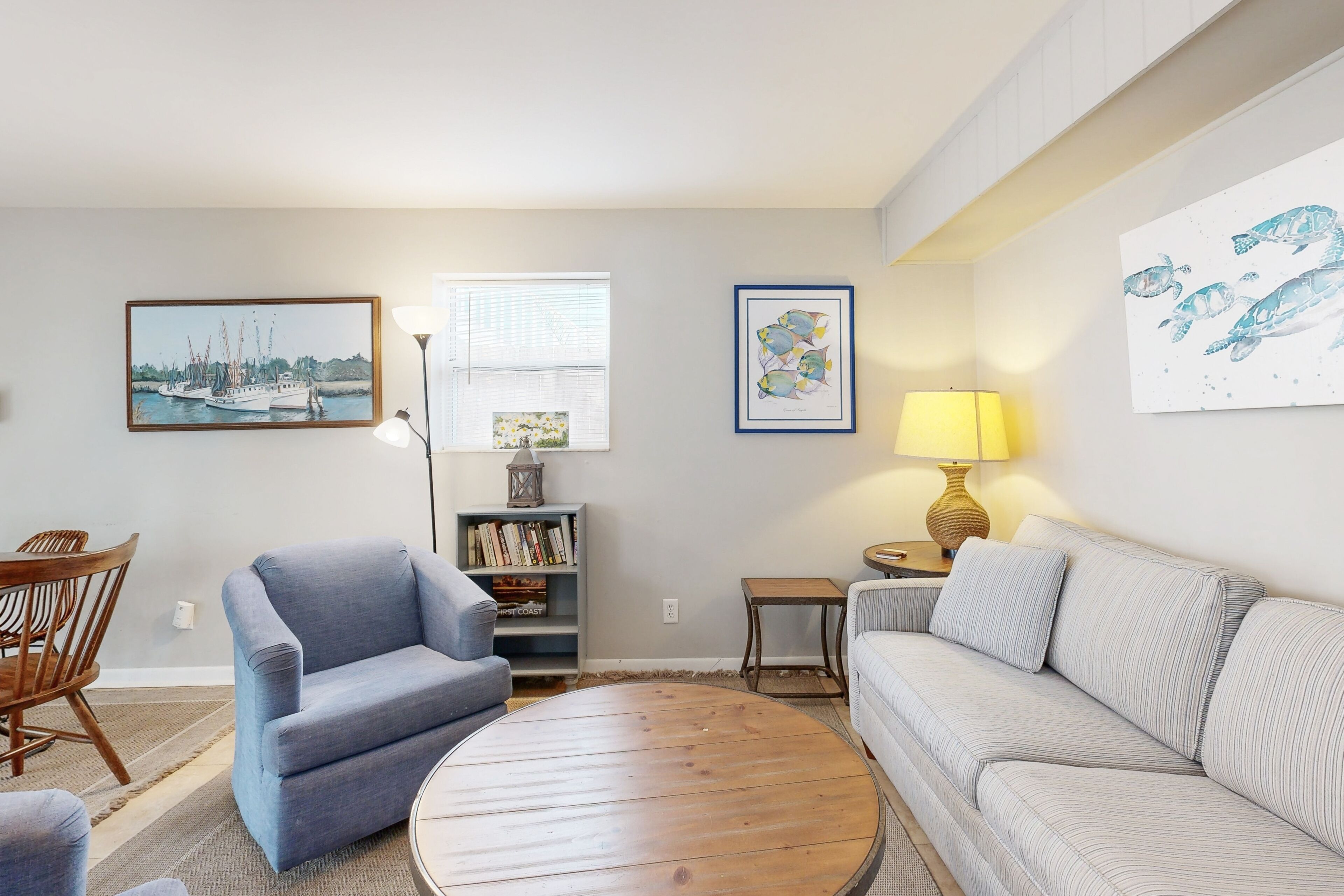 Een woonkamer met een blauwe fauteuil, een witte bank, een ronde houten salontafel, een boekenkast en wanddecoratie met schepen en vissen.