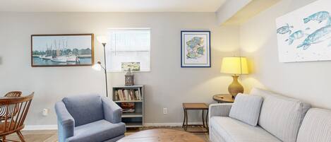 Een woonkamer met een blauwe fauteuil, een witte bank, een ronde houten salontafel, een boekenkast en wanddecoratie met schepen en vissen.