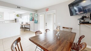 Una cocina moderna con armarios blancos, un comedor con mesa de madera y sillas de mimbre, y un televisor de pantalla plana montado en la pared.