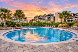 A swimming pool with a curved edge, surrounded by lounge chairs and palm trees, with a backdrop of multi-story buildings. - Ocean-view 4th-floor condo with balcony, outdoor pool & central AC (Fernandina Beach)