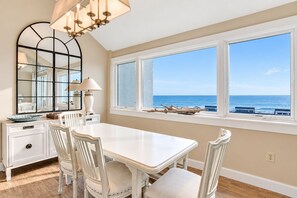 A dining area with a white table and chairs, a large mirror, and a view of the ocean through large windows. - Ocean-view 4th-floor condo with balcony, outdoor pool & central AC (Fernandina Beach)