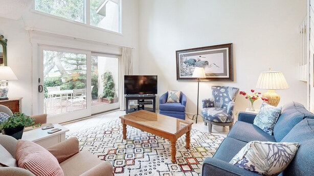 A spacious living room with a large window, a flat-screen TV, a coffee table, sofas, and a patterned rug. - Golf-Course-Front Amelia Island Townhome with Patios, Fast WiFi & Shared Pool (Fernandina Beach)