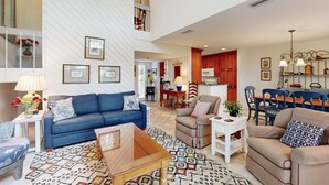 A spacious living area with a blue sofa, beige armchairs, and a wooden coffee table. A dining area with blue chairs and a white table is visible in the background. - Golf-Course-Front Amelia Island Townhome with Patios, Fast WiFi & Shared Pool (Fernandina Beach)