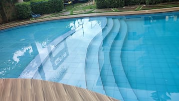 Una piscina con muelle de madera en primer plano, rodeada de vegetación y un edificio al fondo.