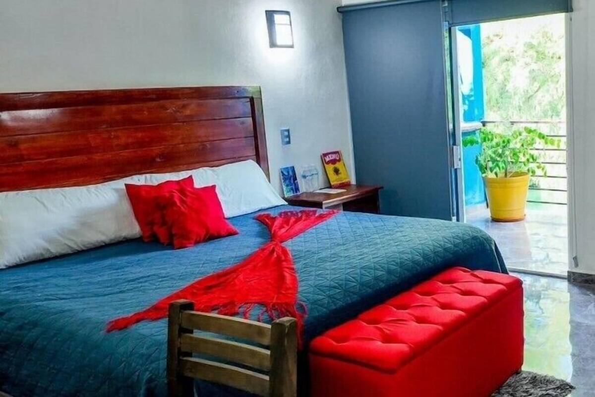 A bedroom with a wooden headboard, a bed with blue and red bedding, a red ottoman, and a potted plant by the window.