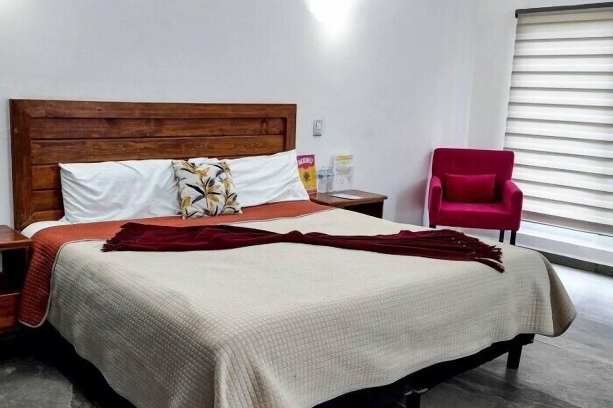 A neatly made bed with a wooden headboard, a red chair, and a window with blinds.