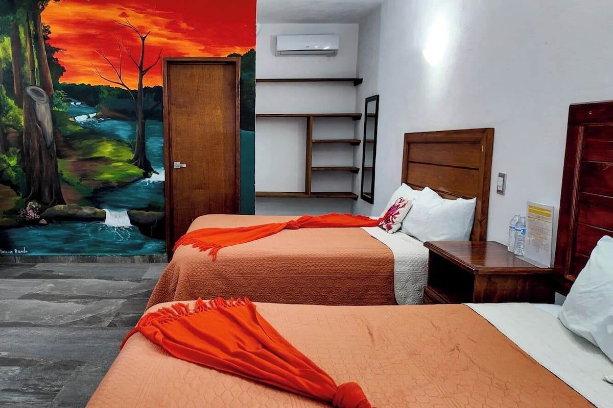 A hotel room with two beds, a mural of a forest and river, a red chair, and a wooden nightstand.