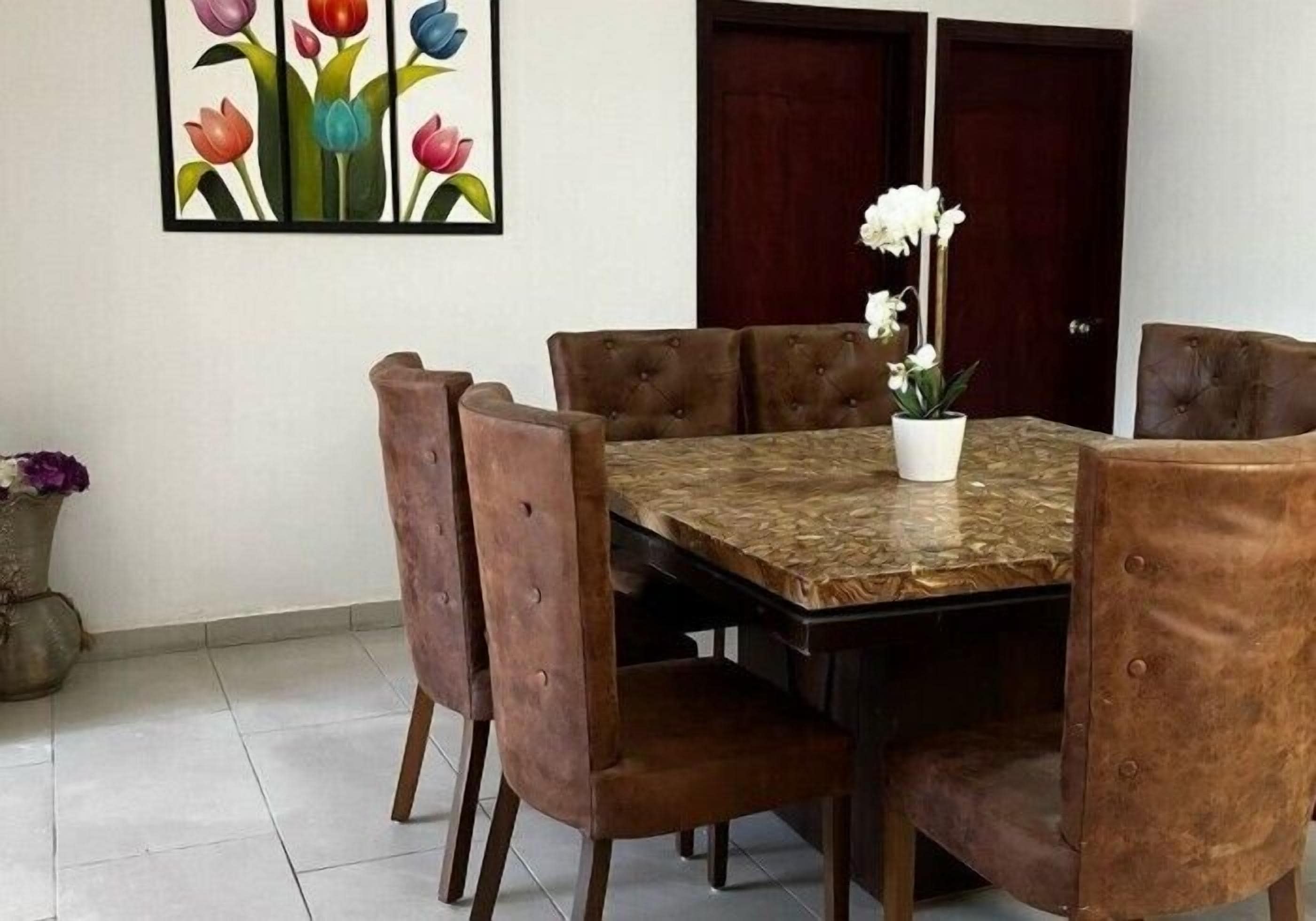 Un comedor con una mesa rectangular rodeada por seis sillas marrones tapizadas. Un florero blanco con flores blancas está sobre la mesa.
