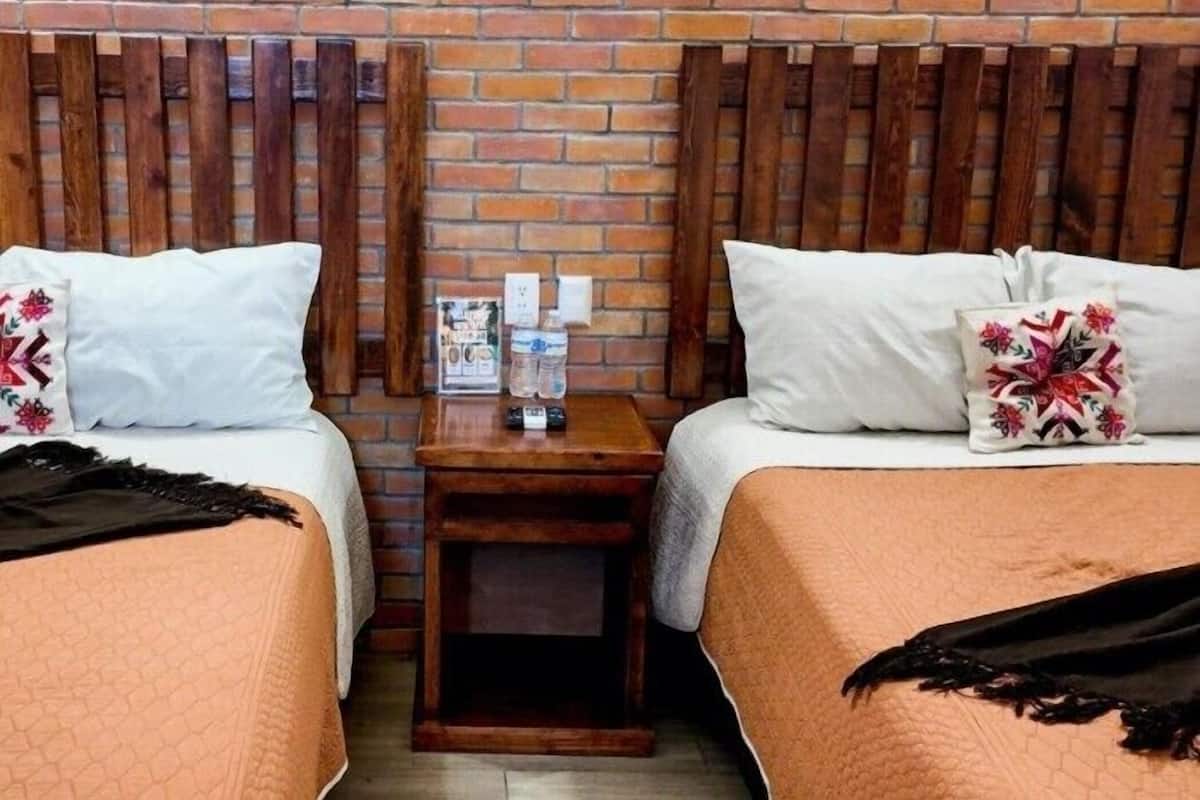 A hotel room with two beds, each with a white pillow and a floral pillow, a wooden headboard, a small wooden nightstand with a bottle and glasses, and a brick wall.