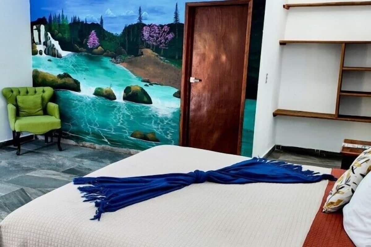 A bedroom with a bed, a green armchair, a mural of a river, and a wooden door.